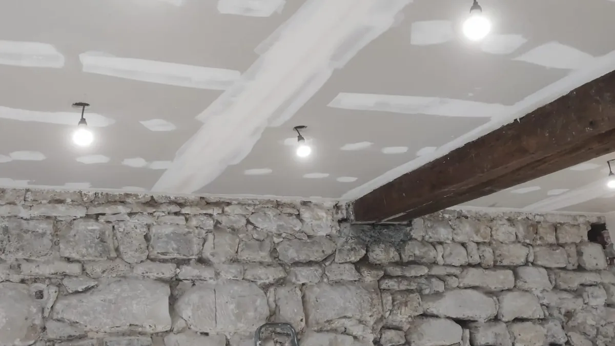 Mur de pierre mis à nu sur lequel repose une poutre en bois, sous un plafond fraîchement plâtré